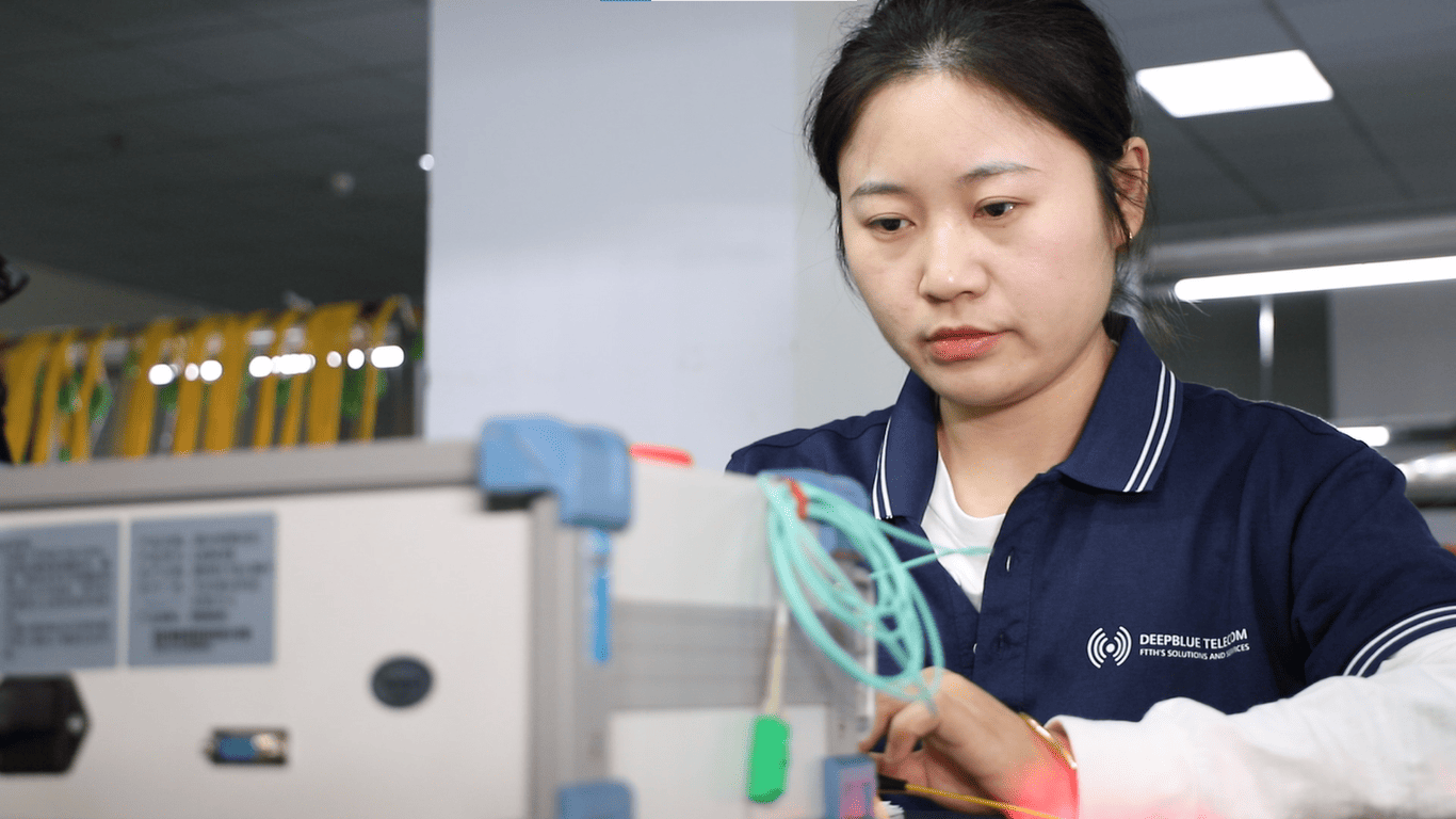 Female technician in a navy polo with DeepBlue Telecom logo, wiring a telecom device in a lab.