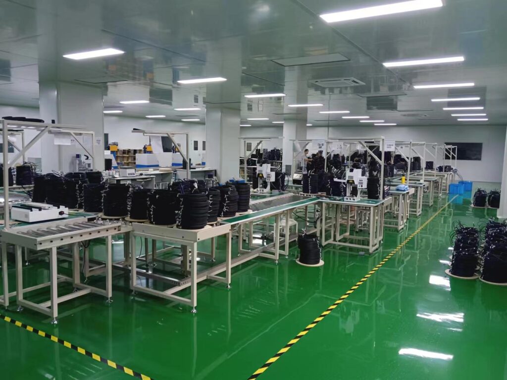 Factory assembly line with racks of black cable reels and workers at workstations in a bright industrial space on a green floor.